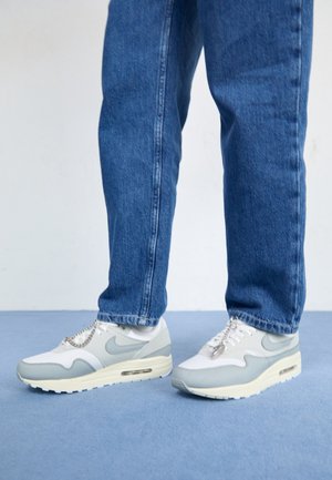 Osoba nosząca niebieskie dżinsy i białoszare buty Nike z metalowym łańcuszkiem na jasnoniebieskiej podłodze przy białej ścianie.