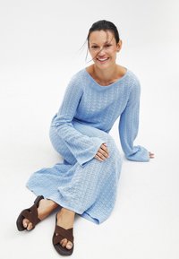 Lichtblauwe gebreide jurk met lange mouwen en uitlopende manchetten, met een gestructureerd patroon. Donkere sandalen met kruislings banden complementeren de outfit.