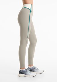 Persoon in beige hoge taille leggings met een teal zijstreep en witte sneakers, zijwaarts staand op een witte achtergrond.