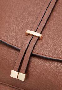 Braune Lederhandtasche mit genarbter Textur, ausgestattet mit zwei Trägern, die durch ein rechteckiges, goldfarbenes Hardware-Accessoire gesichert sind.