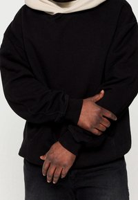 Zwarte hoodie met een beige gevoerde capuchon, dikke manchetten en een losse pasvorm. De textuur is glad met een licht geborstelde binnenkant.