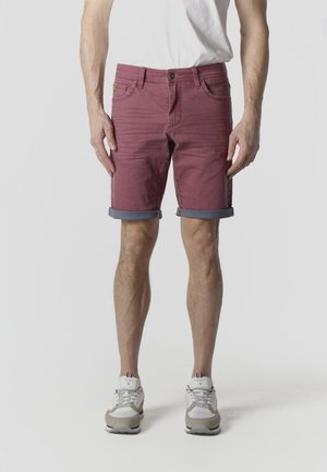 Férfi, aki felhajtott bordó rövidnadrágot, fehér tornacipőt és fehér pólót visel, sima háttér előtt áll.