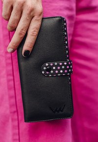 Black leather wallet with a pink and white polka dot strap, featuring a snap closure and embossed logo at the bottom.