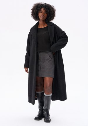 Black long coat in textured fabric, paired with a grey plaid skirt featuring buttons, and knee-high black boots with buckles.