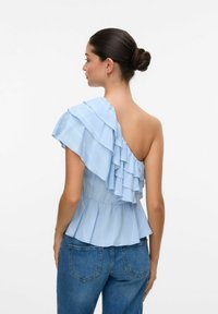 Haut asymétrique bleu clair avec un design à volants sur une épaule, arborant des rayures verticales et une taille cintrée, associé à un jean en denim.