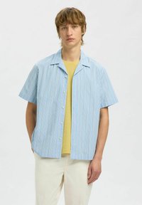 Chemise à manches courtes de couleur bleu clair avec des rayures verticales. Dotée d'un col, d'une fermeture à boutons et d'une coupe décontractée. Associée avec un haut jaune.