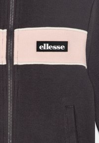 Chaqueta gris oscura con cremallera que presenta una textura acanalada, un panel horizontal rosa con ribete blanco y un parche de logo negro. Bolsillo frontal visible.