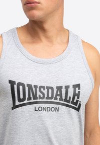Hall kollane ilma varrukateta särk, millel on ümmargune kaelus ja mille esipinnal on silmatorkav must logo, mis ütleb "LONSDALE LONDON".
