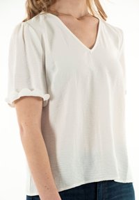 Haut blanc à manches courtes avec un col en V et un ourlet festonné aux manches, fabriqué à partir d'un tissu léger et texturé. Coupe décontractée.