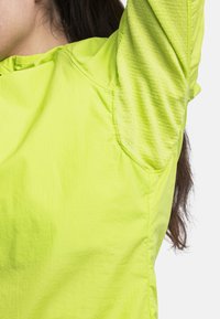 Helle limettengrüne, leichte Sportjacke. Texturiertes Gewebe mit einem Rautenmuster. Verfügt über einen runden Halsausschnitt und verstärkte Schulternähte.