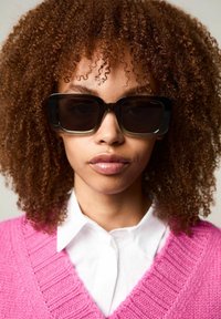 Lunettes de soleil noires surdimensionnées avec des verres foncés, entourées d'un matériau brillant. Le sujet porte une chemise blanche à col sous un pull en laine rose.