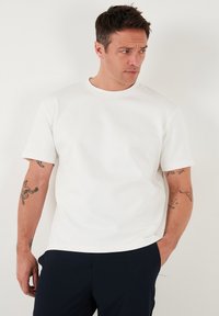 Camiseta blanca de manga corta con un escote redondo, fabricada en un tejido liso y ligeramente elástico. Presenta un corte relajado y sutilezas en las costuras.