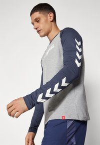 Μπλουζάκι μακρυμάνικο με γκρι σώμα, ναυτικές μανίκες και λευκά μοτίβα ζιγκ-ζαγκ. Σχεδίαση με στρογγυλή λαιμόκοψη, φτιαγμένο από μαλακό ύφασμα. Κόκκινη ετικέτα λογότυπου στο τελείωμα.