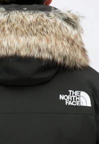 Geacă parka neagră cu glugă căptușită cu blană, având blană falsă de culoare brun deschis și crem. Logo-ul The North Face este afișat proeminent în alb.