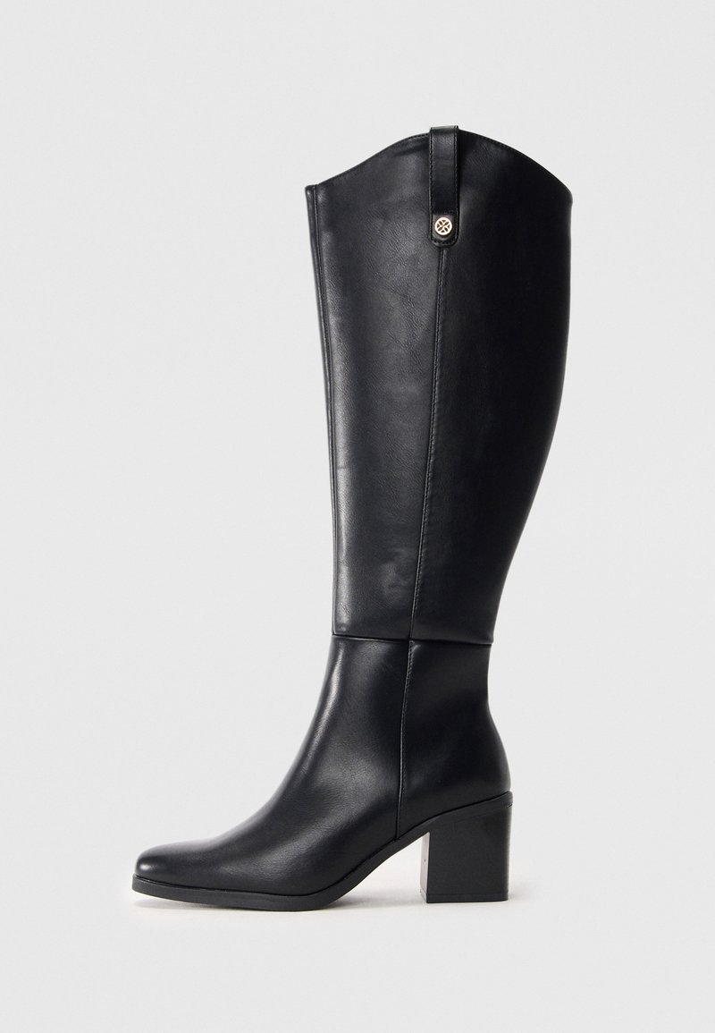 Botte en cuir noir montant jusqu'au genou avec talon carré et languette de traction, ornée d'un petit emblème métallique rond sur la tige extérieure.