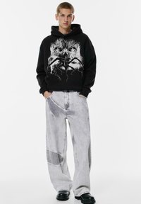 Mladý muž stojí s rukami v kapsách, oblečený v čiernej mikine s kapucňou, na ktorej je grafika lebky a pavúka, v širokých džínsoch s acid-wash úpravou a čiernych čižmách.