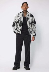 Veste bombardier à motif éclaboussure blanc et noir, fermeture éclair, associée à un débardeur noir et un pantalon noir ample. Des chaussures noires complètent le look.