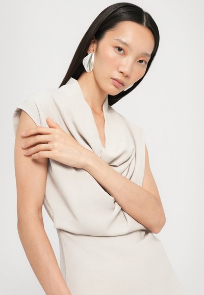 Helle beigefarbene Cowl-Neck-Kleid mit kurzen Ärmeln und drapiertem Stoff. Das Modell trägt große silberne Statement-Ohrringe. Glatte, weiche Textur.