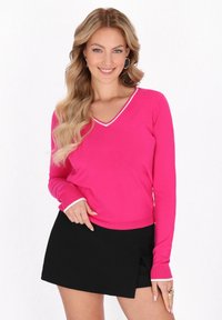 Pulover fucsia cu decolteu în V, cu dantelă albă, mâneci lungi și manșete cusute, asortat cu o fustă neagră tip wrap. Material din tricot fin.