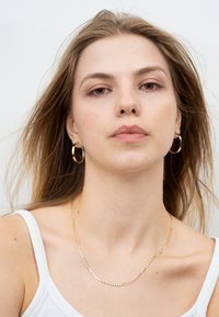 Goldkettchen mit flachem Glieddesign, über einem weißen Tanktop getragen, kombiniert mit goldenen Creolen. Langes, glattes Haar fällt über die Schultern.