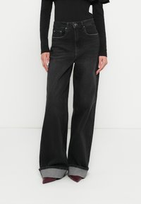 Kõrge vöökohaga mustad teksapüksid laiade jalgadega, millel on kontrastse halli tooniga üleskeeratud äärised. Klassikalised eesmised taskud ja nööpkinnitused.