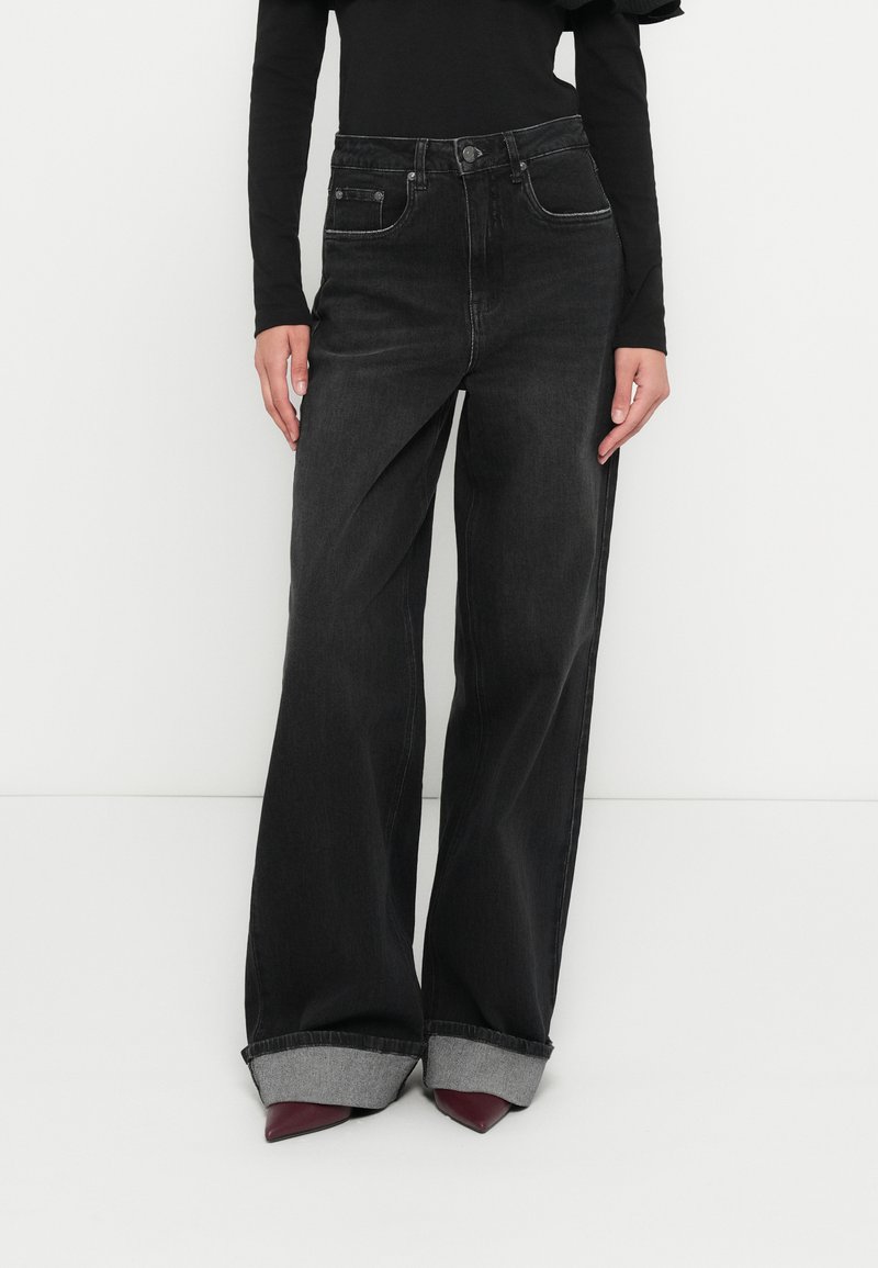 Kõrge vöökohaga mustad teksapüksid laiade jalgadega, millel on kontrastse halli tooniga üleskeeratud äärised. Klassikalised eesmised taskud ja nööpkinnitused.