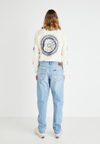 Cămașă crem cu mâneci lungi, având un grafic circular negru pe spate, asortată cu blugi din denim bleumarin deschis. Pantofi albi cu detalii negre de logo.