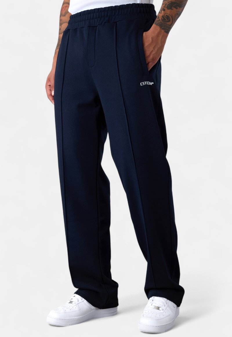 Mann trägt navyblaue, locker sitzende Jogginghosen mit "EST"-Logo auf dem Oberschenkel und weißen Sneakers, steht mit einer Hand in der Tasche.