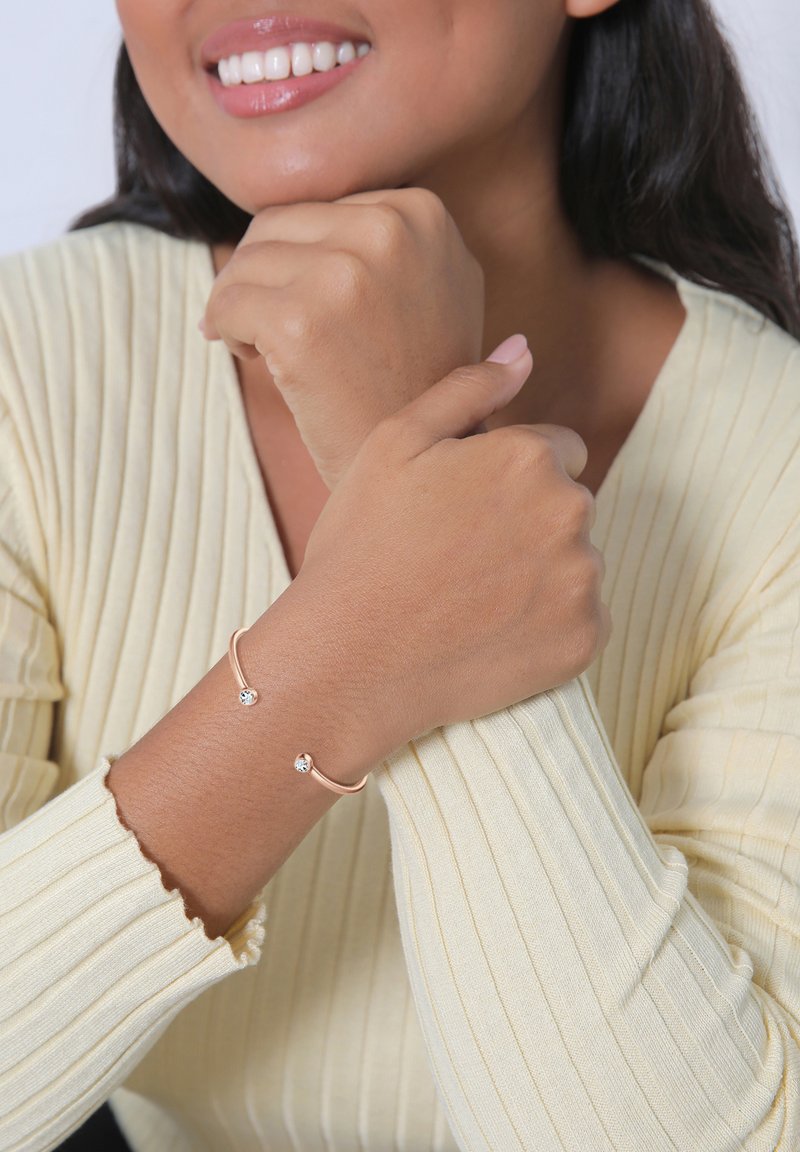 Bracelet en or rose avec deux strass transparents, présentant un design ouvert simple. Porté sur un poignet avec un pull côtelé jaune clair.