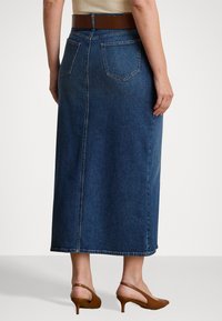 Denim maxiskjørt i mørk blå, med høy midje, to baklommer og en sideglipp. Brukt med brune hæler. Flat, glatt tekstur.