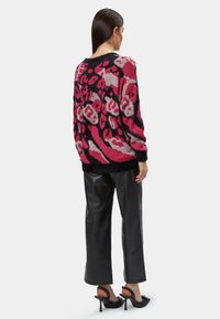 Übergroßer rosa und schwarzer Strickpullover mit abstraktem Leopardenmuster, kombiniert mit schwarzen plissierten Kunstlederhosen und schwarzen High Heels.