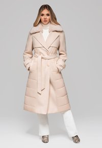 Žena stojící v béžovém kabátu s páskem a kožešinovým límcem, bílém roláku, bílých kalhotách a kotních botách s hadím vzorem.