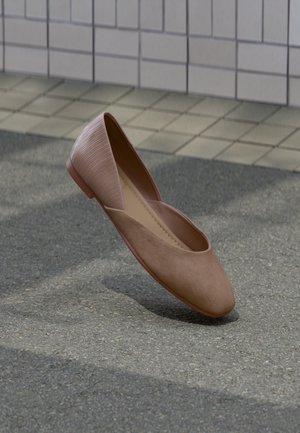 Jednokolorowy beżowy damski lekki but płaski z mieszanymi teksturami unoszący się nad brukowaną powierzchnią w pobliżu ściany pokrytej płytkami.