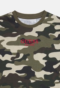 T-shirt i camouflagemönster i gröna och beiga nyanser, med röd broderad text "GUESS EST. 1981" på framsidan, tillverkad av ekologisk bomull.