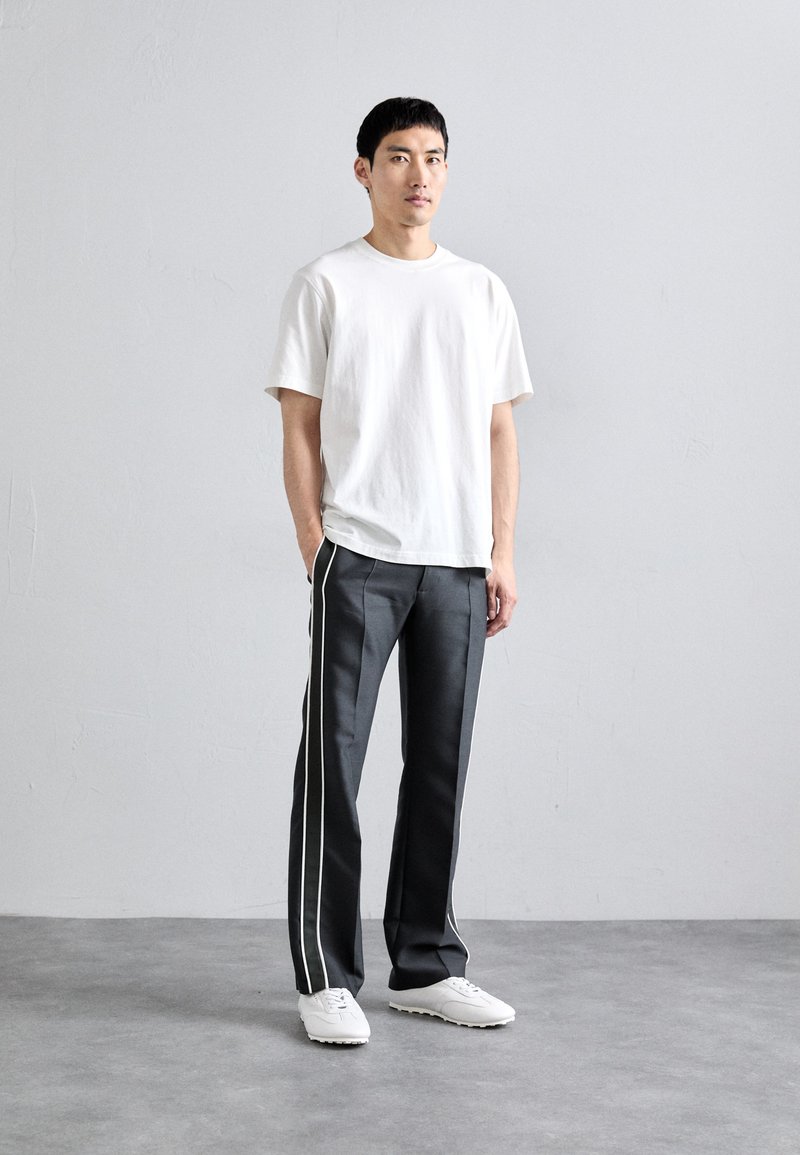 Man staat binnen in een wit T-shirt met korte mouwen, zwarte broek met verticale witte strepen en witte sneakers.