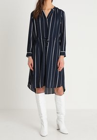 Mörkblå klänning med vita vertikala ränder, långa ärmar, krage, dragsko i midjan och en hi-low fåll. Matchad med vita knähöga stövlar.