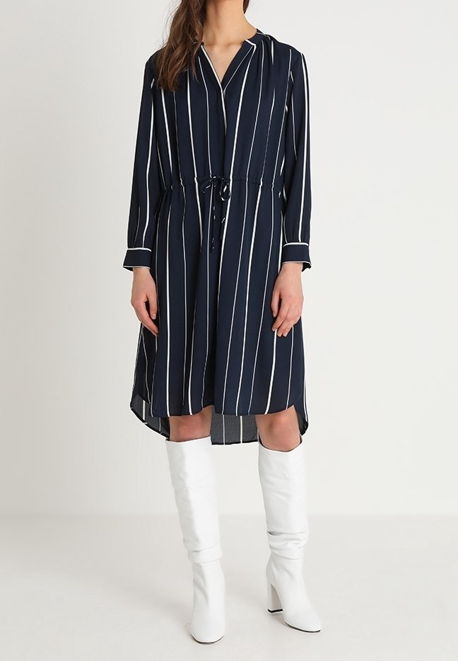 Mörkblå klänning med vita vertikala ränder, långa ärmar, krage, dragsko i midjan och en hi-low fåll. Matchad med vita knähöga stövlar.