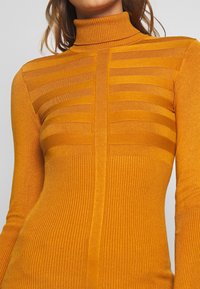Pull en maille côtelée orange avec un col roulé, présentant des motifs de rayures horizontales sur la poitrine, une texture lisse et une silhouette ajustée.