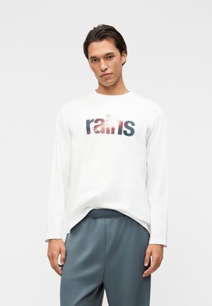 Νεαρός άνδρας που φοράει λευκό πουκάμισο μακρυμάνικο με το κείμενο «rains» και άνετο γκρι παντελόνι, στέκεται μπροστά σε ένα απλό ανοιχτό φόντο.