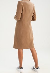 Cappotto in misto lana marrone con maniche lunghe, taglio dritto e cuciture a vista, abbinato a scarpe da ginnastica nero e bianche.