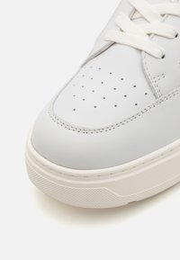 White leather sneaker featuring a rounded toe, perforated details, flat laces, and a textured rubber sole with a light-colored midsole.