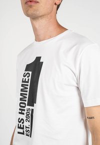 Camiseta de algodón blanca con gran impresión negra "LES HOMMES 1" y "EST. 2005" en el frente. Mangas cortas y cuello redondo.