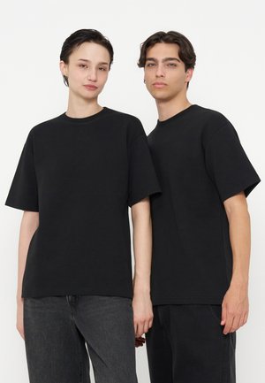 To voksne med kort mørkt hår iført almindelige sorte t-shirts og mørke jeans står side om side mod en hvid baggrund.