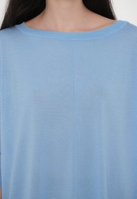 Femme portant un haut tricoté bleu clair avec un large col rond, montrant une partie de son cou et de ses épaules et de longs cheveux foncés.