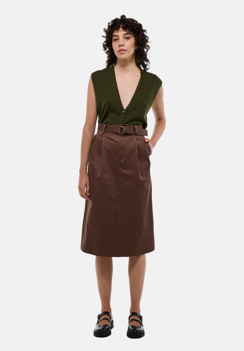 Femme aux cheveux bouclés portant un haut sans manches vert olive, une jupe midi marron ceinturée, et des chaussures Mary Jane noires, debout avec une main dans la poche.