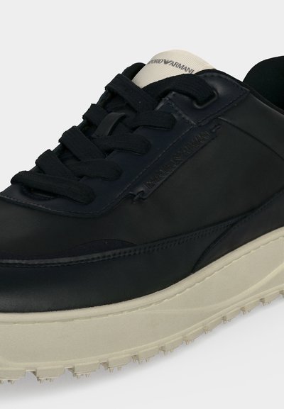 Μαύρο αθλητικό παπούτσι Emporio Armani με παχιά μπεζ σόλα, μαύρα κορδόνια και λογότυπο στη γλώσσα, σε λευκό φόντο.