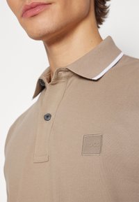 Gros plan d'une personne portant un polo beige avec une bande blanche sur le col et un logo BOSS brodé sur la poitrine.