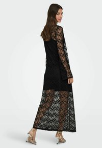 Długa czarna sukienka z koronki z długimi rękawami, zdobiona kwiatowymi wzorami i przejrzystym wyglądem, nałożona na czarną podszewkę.