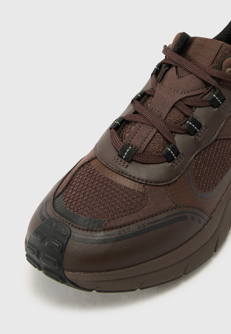 Zapatilla marrón con parte superior de malla texturizada, puntera de goma y sistema de cordones en contraste. Presenta una suela acolchada y acentos suaves.