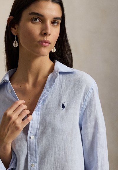 Γυναίκα με μακριά σκούρα μαλλιά, που φοράει πουκάμισο ανοιχτού γαλάζιου χρώματος με μικρό κεντημένο λογότυπο και σκουλαρίκια σε σχήμα κοχυλιού, μπροστά σε ουδέτερο φόντο.